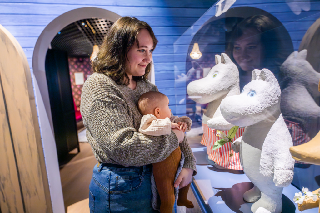 Ihminen pitää vauvaa sylissään ja katsoo Muumihahmoja Muumimuseossa.